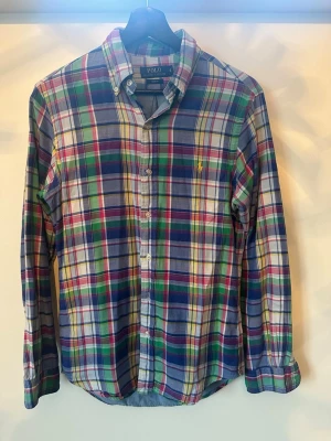 Rutig skjorta från Polo Ralph Lauren - Klassisk rutig skjorta från Polo Ralph Lauren i bomull. Skjortan har knappar framtill, lång ärm och ikonisk broderad logga på bröstet. Färgerna är blå, grön, röd, gul och vit. Perfekt för en avslappnad men ändå stilsäker look.