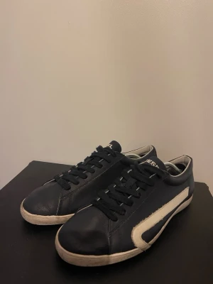 Blåa Diesel sneakers med vit detalj - Snygga svarta sneakers från Diesel med vit sula och vit sidodetalj. Skorna har lågt skaft,blåa skosnören och är tillverkade i slätt läder. Perfekta för dig som gillar en clean och stilren look.