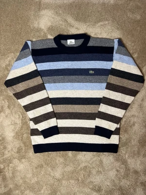 Randig stickad tröja från Lacoste - Snygg stickad tröja från Lacoste med breda horisontella ränder i blått, grått, beige och svart. Tröjan har rund halsringning, långa ärmar och den klassiska gröna krokodilloggan broderad på bröstet. Perfekt för dig som gillar stilrena och tidlösa plagg. Använd fåtal gånger.