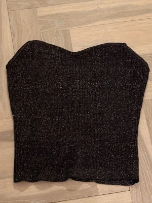 Svart glittrig tubtopp - Snygg svart tubtopp med glittrigt material och ribbad struktur. Toppen är axelbandslös och har en croppad passform som passar perfekt till höga jeans eller kjol. Perfekt för dig som vill sticka ut lite extra. Prislapp kvar
