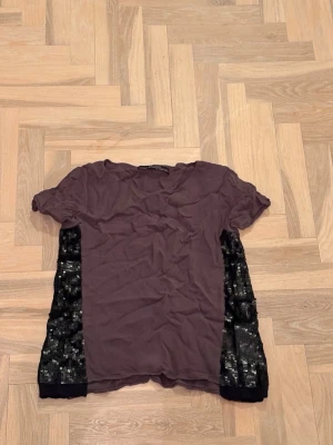 Lila topp med svarta spetsdetaljer - Trendig brun svart topp med korta ärmar och svarta spetsinslag längs sidorna. T-shirten har en avslappnad passform och en klassisk rund halsringning. Perfekt för dig som gillar unika detaljer och en cool look.