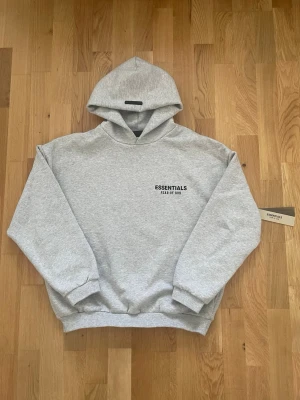 Grå Essentials Fear of God hoodie - Säljer en hoodie från Essentials med en oversized passform, tillverkad i bomull, och som är perfekt för en chill och avslappad stil                                                                           •Färg: Grå                                                                  •Storlek: S                                                              •Typ: Essentials hoodie FW24                                      •Skick: Nytt skick, helt oanvänd