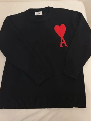 Svart stickad tröja från Ami - Säljer en svart stickad tröja från Ami med deras ikoniska röda hjärta och bokstaven A broderat på bröstet. Tröjan har rund halsringning, ribbade muddar och är gjord i mjukt material som känns skönt mot huden.