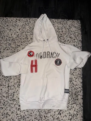Vit Hoodrich hoodie med patchar - Snygg vit hoodie från Hoodrich med stora broderade patchar och logga framtill. Klassisk känguruficka och dragsko i huvan. Perfekt för dig som gillar streetwear och vill sticka ut med detaljer i rött och grått.