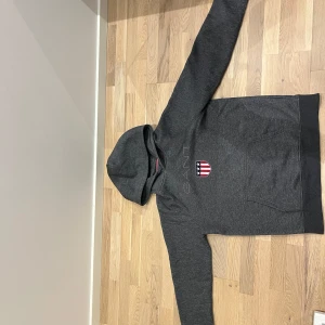 Mörkgrå hoodie från GANT med tryck - Mörkgrå hoodie från GANT med broderad logga och amerikanskinspirerat sköldtryck på bröstet. Klassisk känguruficka, huva och ribbade muddar. Skön och stilren, perfekt till jeans eller joggers.