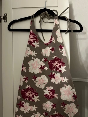 Blommig halterneck linnetopp - Snygg strandtopp med halterneck och knytband i nacken. Toppen har ett grått tyg med stora hibiskusblommor i rosa, vitt och vinrött. Materialet känns som bomull eller bomullsblandning.Perfekt för stranddagar och sommarhäng. ⭐️Halterneck toppen är i mycket bra skick💕