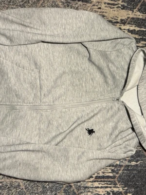 Grå hoodie med dragkedja från Manfinity - Snygg ljusgrå hoodie från Manfinity med dragkedja framtill och svart broderad logga på bröstet. Hoodien har huva och är tillverkad i mjukt, fleeceliknande material på insidan. Perfekt för en avslappnad och sportig stil.