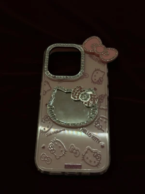 Hello Kitty rosa IPhone 15 PRO mobilskal med glitter - Säljer ett rosa mobilskal med Hello Kitty-design, dekorerat med glittriga stenar runt kameralinsen och på Hello Kitty-bågen. Skalet har upphöjda mönster och en spegel i form av Hello Kittys ansikte. Tillverkat i plast och passar perfekt för dig som gillar söta detaljer. Gäller iPhone 15 PRO. 