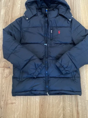 Svart pufferjacka från Ralph Lauren - Säljer en svart pufferjacka från Ralph Lauren med huva och dragkedja framtill. Jackan har en röd broderad logga på bröstet och praktiska fickor med dragkedja. Perfekt för kalla dagar och riktigt skön att bära. Klassisk och stilren design.