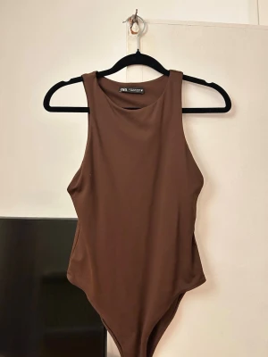 Brun body från Zara - Säljer en stilren brun body från Zara med rund halsringning och ärmlös design. Tillverkad i ett mjukt och stretchigt material som sitter snyggt på kroppen. Perfekt att matcha med jeans eller kjol för en trendig look.