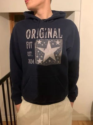 Mörkblå hoodie med stjärntryck - Mörkblå hoodie från Original Fit med stor vit stjärna och texten 'ORIGINAL FIT EST. 2024' på bröstet. Hoodien har huva med dragsko och en klassisk magficka. Perfekt för en avslappnad och cool stil.