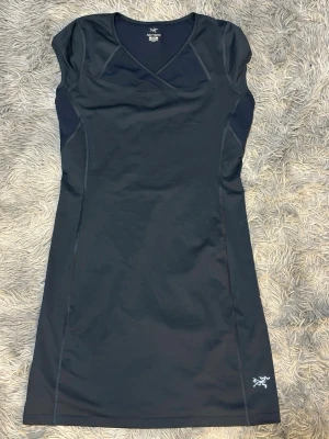 Marinblå sportklänning från Arc'teryx - Snygg marinblå sportklänning från Arc'teryx med v-ringad omlott och korta ärmar. Klänningen är tillverkad i ett syntetmaterial som är perfekt för träning och har en figurnära passform. Diskret logotyp på nederkanten. Perfekt för dig som gillar en aktiv livsstil.