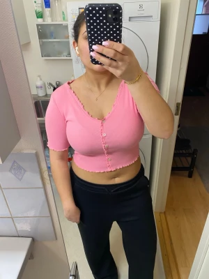 Croppad rosa ribbad topp med knappar - Säljer en croppad rosa topp med ribbad struktur och korta ärmar. Toppen har v-ringning och dekorativa knappar framtill samt volangdetaljer längs kanterna. Perfekt för dig som gillar en färgglad och trendig stil.