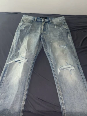 Ljusa slitna jeans Supply & Demand - Säljer ett par ljusblå jeans från Supply & Demand med snygga slitningar och hål på benen. Jeansen har klassisk femficksmodell, raka ben och cool tvättad look. Perfekta för dig som gillar streetwear och avslappnad stil.