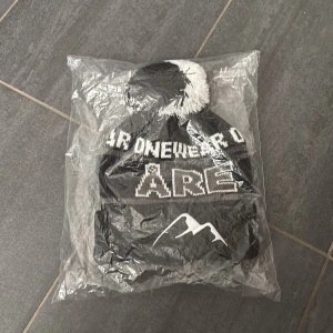 Svart och vit Åre-mössa  - Säljer en svart och vit stickad mössa från Onewear med stor tofs på toppen. Mössan har texten 'ÅRE' och ett stiliserat bergmotiv framtill. Perfekt för kalla dagar och vintersäsongen. Jag van den på en intervju med Bruce lee men har inte öppnat den.