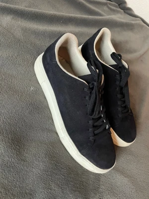 Marinblå selected homme skor - Marinblå selected homme i mocka med vita sulor och svarta skosnören. Insidan är ljus och skon har en klassisk, stilren design som passar till det mesta. Perfekta för dig som gillar en clean look. Skorna är använda en del och har sina nackdelar, har även en liten slitning vid hälen på insidan av skon.