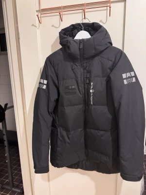 Svart dunjacka från Sail Racing - Säljer en svart dunjacka från Sail Racing med huva och dragkedja. Jackan har loggor på ärmarna och bröstet samt en bröstficka med dragkedja. Perfekt för kalla vinterdagar och riktigt snygg streetstil.