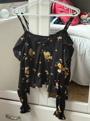 Blommig svart topp från H&M Divided - Svart blommig topp från H&M Divided med smala axelband och långa ärmar med volangdetaljer. Toppen har ett gult och vitt blommönster och en croppad passform. Perfekt för dig som gillar trendiga och feminina plagg.