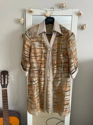 Beige mönstrad skjorta  - Unik beige skjorta från McBorrow med abstrakt mönster i bruna och orange toner. Skjortan har klassisk krage, knappar framtill och korta ärmar med uppvik. Den är genomskinlig i vissa rutor ,man ser på bild 6.den är lång nästan ovan på knäna om man är 163cm .Perfekt för dig som vill sticka ut med en cool vintage vibe.