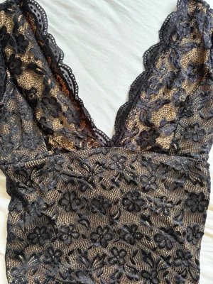 Svart spetstopp med djup urringning - Snygg svart spets topp med blommigt mönster och djup v-ringning både fram och bak. Bodyn har transparent spets över beige tyg och dekorativa kanter. Perfekt för dig som gillar feminina detaljer och trendig design.