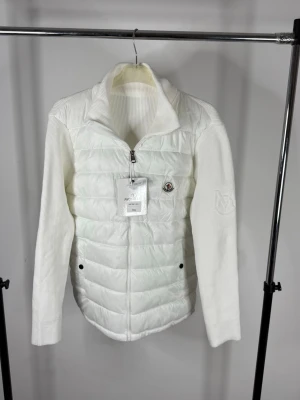 Moncler cardigan  - Säljer en riktigt snygg och exklusiv vit Moncler hybrid-cardigan i nyskick. Denna modell kombinerar den klassiska quiltade dunvästen på framsidan med lyxigt stickade ärmar och rygg – perfekt för både vår, höst eller som ett mellanlager under kallare dagar. Detaljer: • Märke: Moncler • Färg: Vit • Storlek: 1XL (Se bild på tagg för referens) • Skick: 5/5 – Helt ny med alla tags kvar! • Design: Quiltad framsida med dragkedja, ribbstickade ärmar och krage. Den klassiska Moncler-loggan pryder bröstet