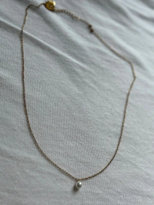 Edblad Guldhalsband med pärlhänge - Snyggt och minimalistiskt halsband i guldton med en liten vit pärla som hänge. Kedjan är tunn och elegant, perfekt för att ge en stilren touch till din outfit. Passar dig som gillar klassiska accessoarer med en modern twist.