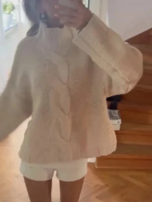 Beige kabelstickad tröja med hög krage - En oversized beige stickad tröja med bred kabelstickning framtill och ribbade detaljer vid krage, ärmslut och nederkant. Tröjan har hög krage och långa ärmar, perfekt för att hålla sig varm och snygg under kyliga dagar.