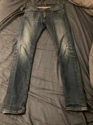 Blå slitna jeans från Lee - Säljer ett par blå jeans från Lee med slitningar och en mindre reva på höger lår. Jeansen har klassisk femficksmodell, raka ben och snyggt tvättad denim. Perfekta för en avslappnad och trendig look.   Storlek: W31 L34