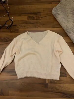 Ljusbeige stickad V-ringad tröja - Mysig ljusbeige stickad tröja med V-ringning och långa ärmar. Tröjan har ribbstickade muddar vid ärmslut och nederkant, perfekt för lager på lager. Enkel och stilren design som passar till det mesta.