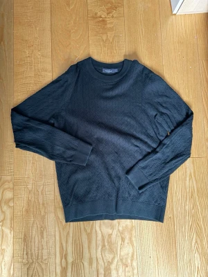 Mörkgrön stickad tröja  - Svart långärmad tröja från Dressmann med diskret stickat pilmönster över hela plagget. Tröjan har rund halsringning och ribbade muddar vid ärmslut och nederkant. Perfekt för en stilren och enkel look.