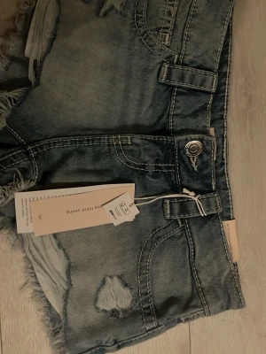 Blå slitna jeansshorts från Gina Tricot - Säljer ett par blå denimshorts från Gina Tricot med låg midja, slitna detaljer och fransiga benslut. Klassisk femficksmodell med knapp och dragkedja. Perfekta för sommaren och ger en avslappnad streetstyle-look.