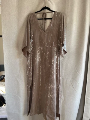 Glittrig beige/grå klänning med v-ringning - Snygg beige/grå balklänning med glittriga paljetter och v-ringning. Klänningen till mina fotknölar (jag är ca 170), vida ärmar och knytband i nacken. Perfekt för dig som vill sticka ut med en elegant look. Lite stor på mig som brukar ha M så skulle säga att de är en L