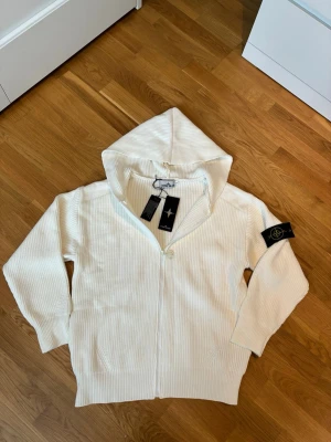 Stone Island zip-up hoodie - Snygg beige zip-up hoodie från Stone Island i ribbstickad bomull. Dragkedja framtill, huva och klassisk logotyp-patch på ärmen. Perfekt för en clean och stilren look. Diskret men ändå med tydlig streetwear-vibe.