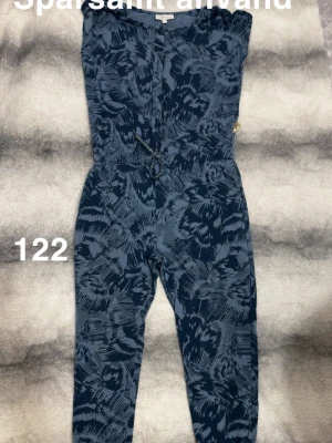 Mönstrad blå jumpsuit med dragsko - Snygg blå jumpsuit med abstrakt mönster och dragsko i midjan. Ärmlös design och långa ben, perfekt för en trendig och avslappnad look. 🌸 Sparsamt använd. Storlek 122 