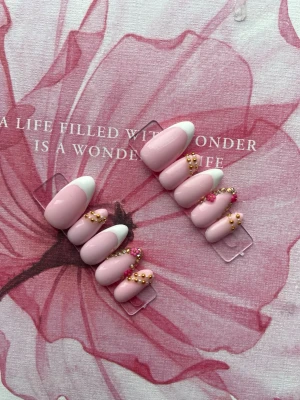 Rosa press-on naglar med pärldekor - Snygga press-on naglar i ljusrosa med vit fransk tipp och dekorerade med små guld- och rosa pärlor. Naglarna har en oval form och ger en elegant look. Perfekt för dig som vill ha en trendig och unik nageldesign utan att gå till salong.