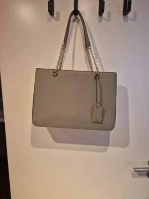 Grå handväska från DKNY med kedjedetaljer - Snygg grå handväska från DKNY i stilren design. Väskan har silverfärgade kedjehandtag och ett matchande bag tag med DKNY-logga. Tillverkad i ett tåligt material med struktur och har plats för allt du behöver. Perfekt accessoar för en trendig look.