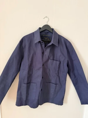 Blå overshirt från D'once De La Sense - Snygg blå overshirt från D'once De La Sense med klassisk krage med  fickor framtill! Nästintill oanvänd. Tillverkad i ett robust tyg som ger en stilren och avslappnad look. Perfekt att bära över en skjorta eller t-shirt.