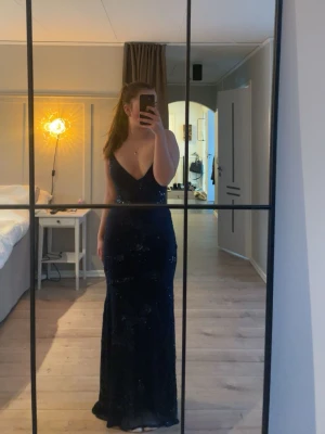 Marinblå långklänning med öppen rygg - Säljer en elegant marinblå långklänning med v-ringad front och öppen rygg med snörning. Klänningen har glittriga detaljer och smala axelband som korsas i ryggen. Perfekt för bal eller fest. Klänningen har spruckit lite u det yttre tyget kan skicka bild vid intresse men det sys lätt ihop 