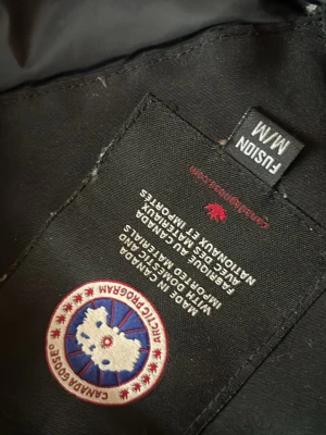 Svart dunjacka från Canada Goose - Säljer en svart dunjacka från Canada Goose med klassisk patch på ärmen och huva med pälskant. Jackan har dragkedja framtill och är tillverkad i slitstarkt material, perfekt för kalla vinterdagar. Stilsäker och varm jacka som håller dig skyddad mot kyla.