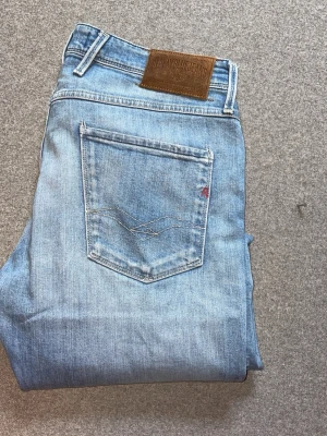 Ljusblå Replay jeans med skinnpatch - Säljer ett par ljusblå jeans från Replay med klassisk femficksmodell och snyggt slitna detaljer. Jeansen har en brun skinnpatch med Replay-logga bak i midjan och raka ben. Materialet är mjukt denim i bomull, perfekt för en avslappnad stil.