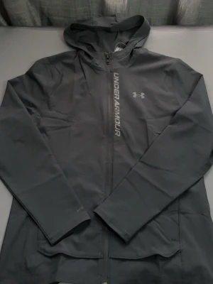 Grå vindjacka från Under Armour - Sportig grå vindjacka från Under Armour med huva och dragkedja framtill. Jackan har lång ärm, tryckt logga och text på bröstet. Perfekt för dig som gillar en clean och funktionell stil.