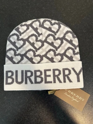 Gråvit Burberry mössa med logotyp - Snygg gråvit mössa Burberry med tydlig logotyp framtill och mönster av Burberrys klassiska monogram. Mössan är stickad och har en uppvikt kant. Perfekt accessoar för att lyfta din stil under kalla dagar.