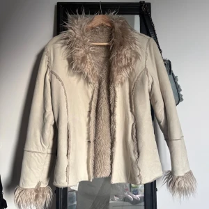 Beige/vit pälsjacka med fluffig krage - Stilren beige pälsjacka med fluffig, långhårig fuskpäls vid krage, ärmslut och insida. Jackan har en figurnära passform och stängs med knappar framtill. Perfekt för dig som vill ha en trendig vårlook!🌺