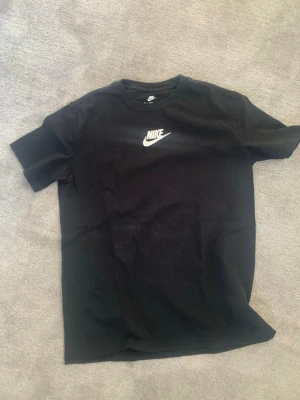 Svart Nike t-shirt med logga - En svart t-shirt från Nike med klassisk vit logga på bröstet. T-shirten har rund hals och korta ärmar, perfekt för en sportig och avslappnad stil. Tillverkad i mjuk bomull som känns skön mot huden.