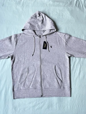 Grå hoodie från Polo Ralph Lauren - Snygg ljusgrå hoodie med dragkedja från Polo Ralph Lauren. Klassisk design med huva, dragsnören och två fickor framtill. Broderad logga på bröstet och mjukt bomullsmaterial. Perfekt för en chill och stilren look.