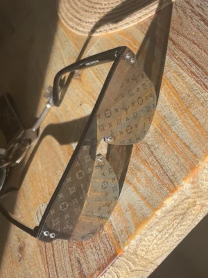 Louis Vuitton svarta solglasögon - Snygga svarta solglasögon från Louis Vuitton med stora, rektangulära glas och klassiskt LV-monogram över hela linsen. Bågarna är svarta och har en modern, kantig design. Perfekta för dig som vill sticka ut med en lyxig accessoar.