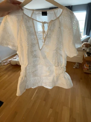 Vit broderad blus med knytning från Gina - Superfin vit blus från Gina Tricot med broderade detaljer och hålmönster. Blusen har korta ärmar, v-ringning med knytband och resår i midjan för en snygg passform. Perfekt till jeans eller kjol för en fräsch look.