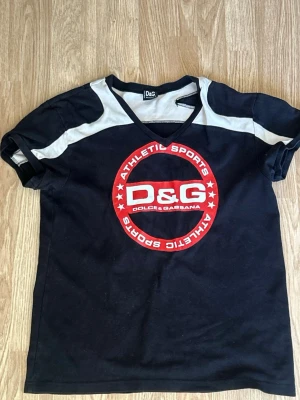 Svart D&G t-shirt med sportig look - Cool svart t-shirt från Dolce & Gabbana med vit detalj på axlarna och stort rött D&G-tryck framtill. V-ringad modell med sportig känsla, perfekt för en avslappnad streetstyle. Skön passform och klassisk kortärmad design.