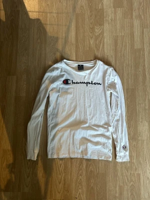 Vit långärmad tröja från Champion - Vit långärmad tröja från Champion med rund halsringning och stort broderat logotyptryck på bröstet. Tillverkad i mjuk bomull som känns skön mot huden. Perfekt för en avslappnad och sportig stil.