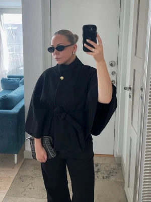 Svart poncho kappa med knytband - Stilren svart poncho-kappa med vida ärmar, knytband i midjan och en dekorativ guldig knapp vid halsen. Kappan har en elegant och modern siluett, perfekt för lager-på-lager och att lyfta din outfit. Varan är använd ett fåtal gånger så den är i ett väldigt bra skick!